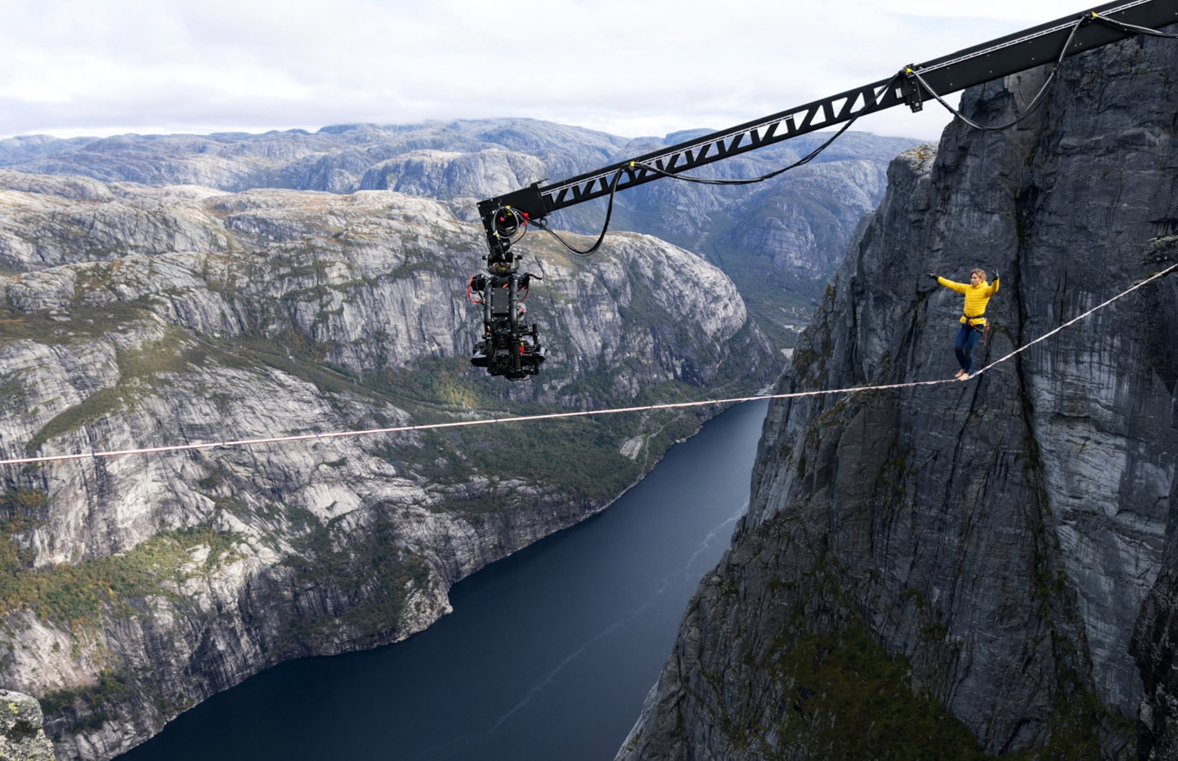 A person in a yellow jacket walking across a rope spread over a valley, with a large camera on a crane recording her.