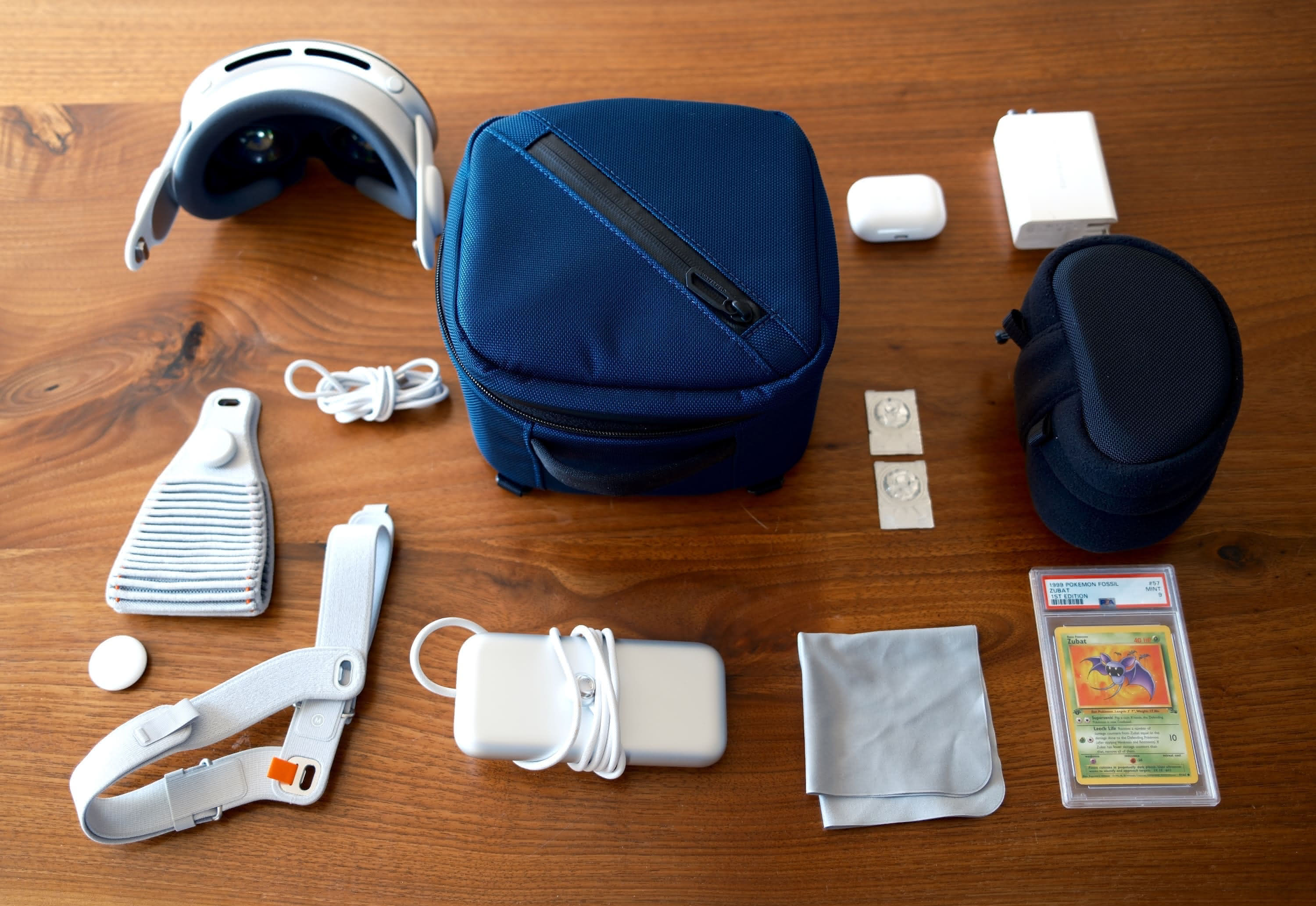 The case on a table showing the items it can carry, including the Vision Pro, a USB-C cable, the two included headbands, an AirTag, the battery, AirPods Pro, a polishing cloth, a PSA 9 first edition Zubat Pokémon card, the inner fleece case, two contact lenses, and a charging brick.