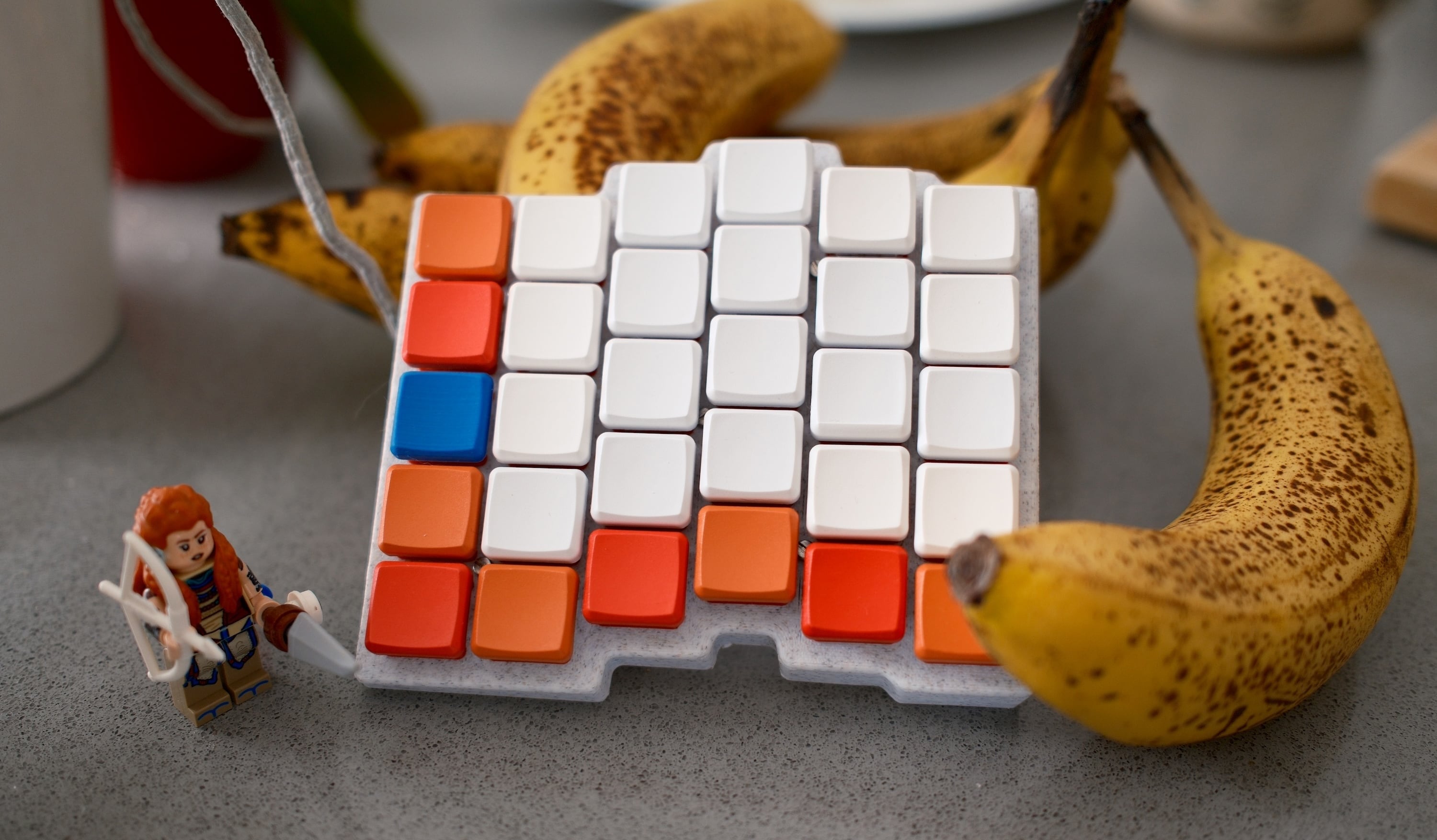 The Caldera keyboard on a kitchen island with bananas around it and a LEGO figurine of Aloy from Horizon Zero Dawn